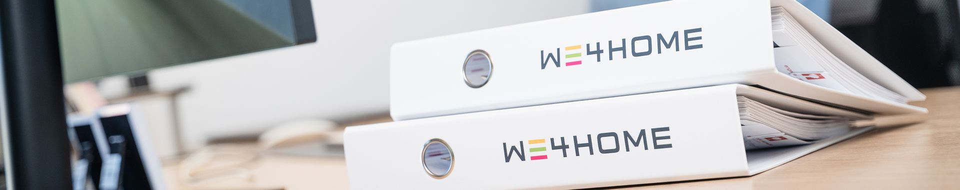 Weiße Ordner mit WE4HOME-Logo auf einem Schreibtisch.